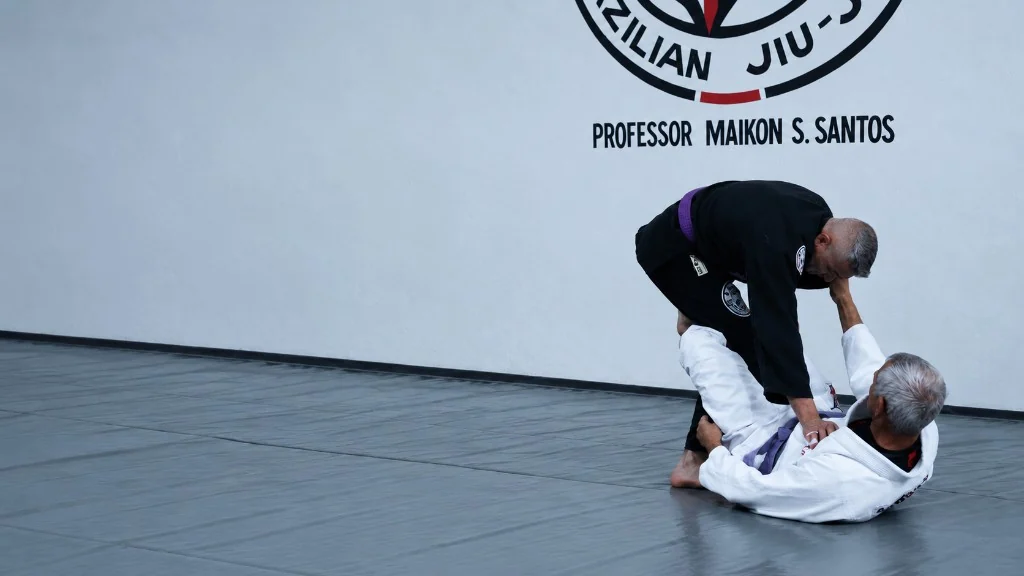 Dois atletas da Aslam BJJ no tatame. Um de kimono preto aplica a posição no outro, de kimono branco